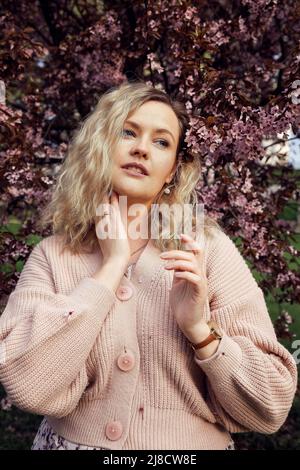 Portrait de belle femme blonde cheveux avec un maquillage doux et des boucles parfaites posant sur fond lilas fleur dans le gilet tricoté rose. Mode Banque D'Images