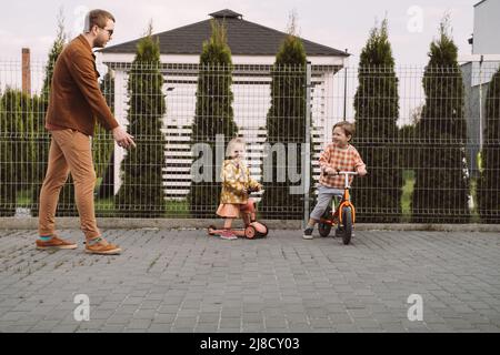 Enfants jouant à la course de vélo et de scooter dans la cour de la maison. Sœur et frère à cheval rapide autour de la maison privée. Concours de frères et sœurs de famille. Enfants Banque D'Images