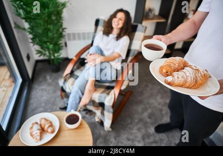 Les hommes tiennent une tasse de café et des croissants tandis que la femme est assise sur un fond flou. Couple amoureux en appréciant le petit déjeuner dans une maison confortable. Concentrez-vous sur les mains masculines avec des pâtisseries et des boissons. Banque D'Images