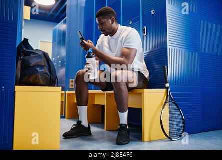 Portrait complet d'un sportif noir musclé utilisant un smartphone dans un vestiaire avec une raquette de tennis en premier plan, espace de copie Banque D'Images