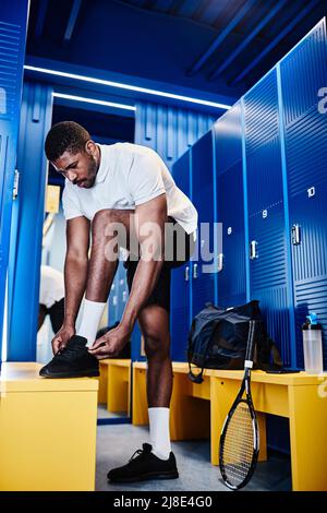 Portrait vertical complet d'un sportif noir qui noue des lacets tout en se changeant dans le vestiaire pour se préparer à l'entraînement au tennis Banque D'Images