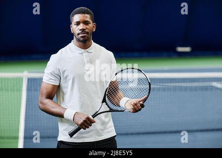 Portrait à la taille haute d'un joueur de tennis afro-américain regardant l'appareil photo tout en posant en confiance avec une raquette sur un court intérieur, espace de copie Banque D'Images