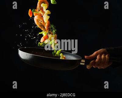 Crevettes tigrées avec des tranches de citron, des légumes et des herbes dans une poêle à frire. Nourriture dans un vol gelé sur fond noir. Le processus de cuisson des fruits de mer wi Banque D'Images