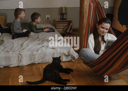 Les enfants jouent à la station de jeux pendant que la mère lit le livre dans le hamac. Famille avec chat noir comme animal de compagnie. Frères jouant à des jeux vidéo sur le lit. Famille Banque D'Images
