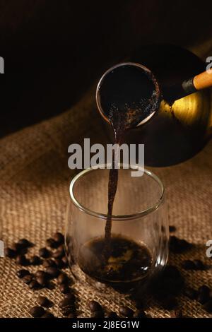 Le café espresso fraîchement préparé est versé d'une glace dans une tasse en verre transparent sur un fond sombre de toile de fond avec des grains de café Banque D'Images