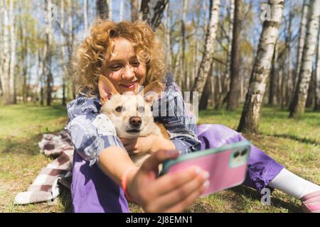 Gros plan d'une joyeuse belle fille caucasienne aux cheveux bouclés, assise sur l'herbe dans un parc tenant un téléphone portable, prenant une photo de son chien et d'elle. Photo de haute qualité Banque D'Images