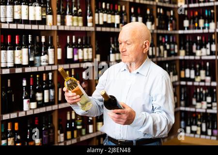 Un homme âgé sérieux choisit entre le vin rouge et le vin blanc dans un magasin de spiritueux Banque D'Images