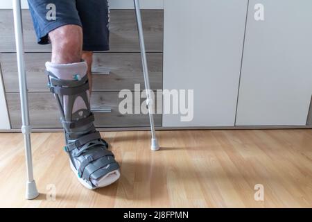 L'homme européen après l'opération de rupture de tendon d'Achille est de retour à la maison avec la chaussure de physiothérapie spéciale de botte de lune et des béquilles pour la récupération à la maison contre Banque D'Images