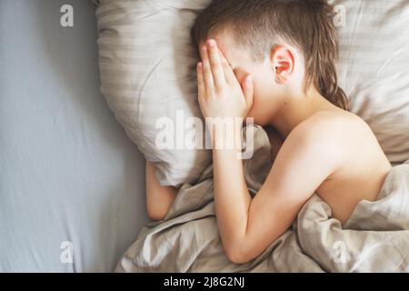 le garçon dorme dans le lit. l'enfant est allongé sur un oreiller et couvre son visage avec ses mains Banque D'Images