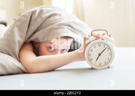 garçon allongé sur le lit sous une couverture et le réveil de l'arrêt le matin. main de childs se levant pour le réveil pour l'éteindre. Banque D'Images