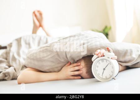 garçon allongé sur le lit sous une couverture et le réveil de l'arrêt le matin. main de childs se levant pour le réveil pour l'éteindre. Banque D'Images