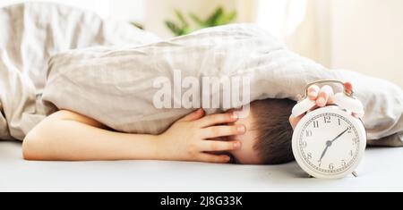 un garçon allongé sur le lit sous une couverture et un réveil le matin. un petit coup de main pour les enfants qui se met à la disposition du réveil pour l'éteindre. bannière Banque D'Images