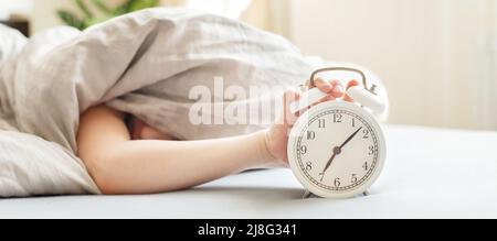 un garçon allongé sur le lit sous une couverture et un réveil le matin. un petit coup de main pour les enfants qui se met à la disposition du réveil pour l'éteindre. bannière Banque D'Images