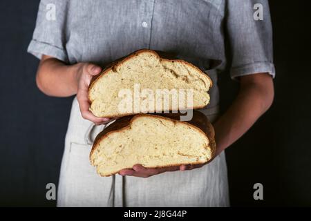 Les mains des femmes tenant une pâte à base de levain fraîchement cuite artisanent le pain coupé en tranches de demi. Maison avec farine de blé et entrée. Style rustique sombre. Banque D'Images