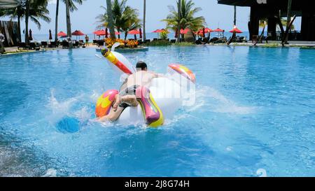 Joyeux drôle homme avec succès sauter sur la licorne gonflable flottant dans la piscine de l'hôtel tropical pour faire du vélo. Fou touriste mâle ont du plaisir sur géant Banque D'Images