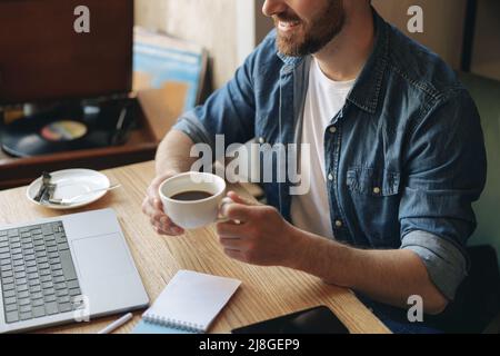 Tasse blanche de café noir dans la main d'un homme. Homme caucasien dans un café travaillant avec un ordinateur portable. Repos. Banque D'Images