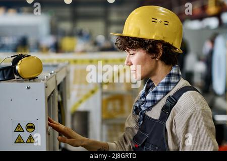 Jeune femme ingénieur ou technicien en vêtements de travail et casque de protection vérifiant l'équipement industriel ou la machine pendant le travail en usine Banque D'Images