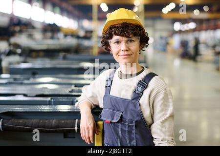 Jeune femme heureuse travailleuse d'usine moderne portant un chandail gris, une combinaison bleue et un casque de sécurité jaune debout près d'un nouvel équipement industriel Banque D'Images