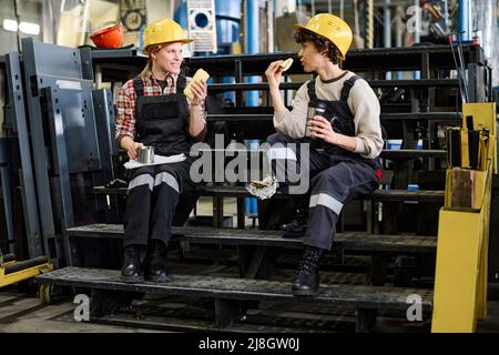 Deux jeunes ingénieurs discutent en étant assis dans l'escalier de l'entrepôt de distribution et en prenant des sandwiches avec du thé pour le déjeuner Banque D'Images