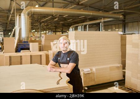 Portrait d'un jeune travailleur dans une combinaison regardant la caméra se tenant à l'entrepôt près des produits en bois Banque D'Images