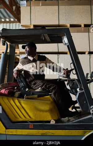 Conducteur de chariot élévateur africain assis dans le chargeur et conduisant pendant le travail à l'usine industrielle Banque D'Images