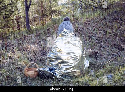 La femme est perdue et s'assoit dans la forêt sauvage par temps froid, en utilisant une couverture de secours d'urgence pour prévenir l'hypothermie et la perte de chaleur corporelle. Banque D'Images