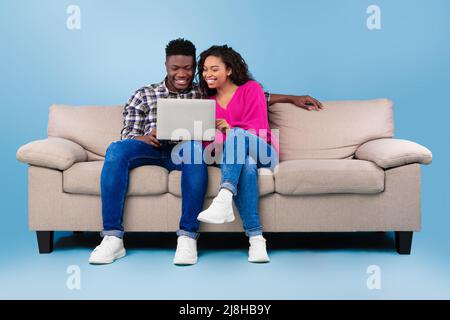 Joyeux jeune couple noir utilisant un ordinateur portable tout en étant assis sur un canapé sur fond bleu studio, pleine longueur Banque D'Images
