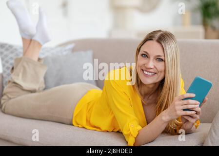 Jolie jeune femme blonde blonde de couleur européenne souriante vêtu de vêtements jaunes avec un smartphone reposant sur un canapé Banque D'Images