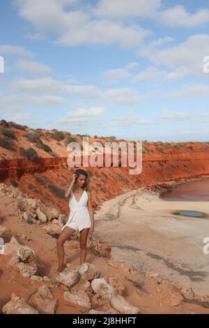 Jolie jeune femme blonde en robe blanche sur la falaise avec plage de sable rouge sur la côte ouest de l'Australie Banque D'Images