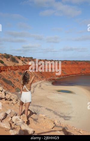 Portrait arrière d'une jolie jeune femme blonde en robe blanche sur une falaise avec plage de sable rouge sur la côte ouest de l'Australie Banque D'Images