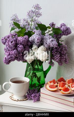 Un bouquet parfumé de lilas colorés est placé dans un vase en verre vert sur fond blanc. À côté d'une tasse de thé chaud et de ​​grapefruit tranché. Banque D'Images