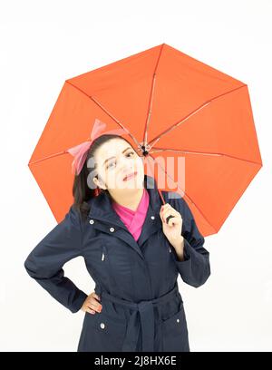 jolie femme asiatique blanche, portant un trench-coat bleu et un chemisier rose, heureuse et satisfaite de tenir un joli parapluie rouge d'une main, sur fond blanc Banque D'Images