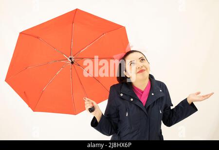 jolie femme asiatique blanche debout, portant un trench-coat bleu et un chemisier rose, notant avec joie qu'elle a cessé de pleuvoir, sous un joli parapluie rouge Banque D'Images