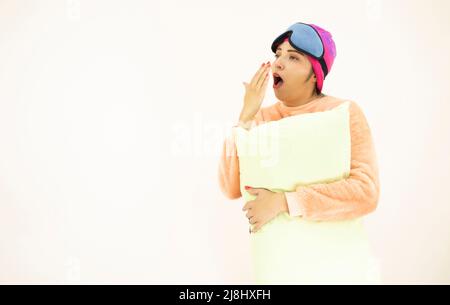 belle jeune femme blanche asiatique portant un pyjama rose, un bonnet de nuit et un masque de sommeil, bâillonner debout et tenant un oreiller, sur fond blanc Banque D'Images