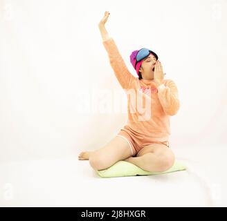 belle jeune femme asiatique fatiguée portant un pyjama rose, bâchant et étirant un bras tout en se reposant sur le sol, sur fond blanc Banque D'Images