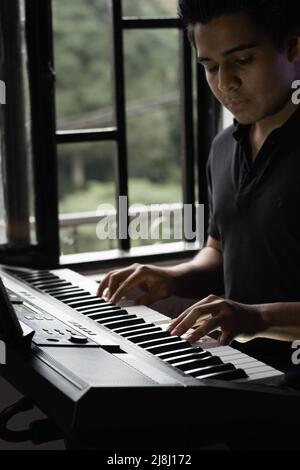 jeune latin jouant du piano électrique ou synthétiseur dans sa maison à côté de sa fenêtre, très concentré, il utilise la musique pour se détendre et penser à h Banque D'Images