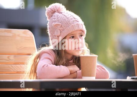 Portrait d'une petite fille mignonne dans un chapeau rose assis seul au café de rue buvant du thé de la tasse en papier Banque D'Images