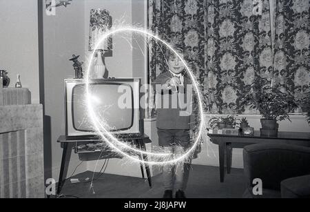 1970s, historique, un jeune garçon de choooboy jouant à l'intérieur dans un salon avec un feu de cheminée, créant un cercle de lumière brûlante, Angleterre, Royaume-Uni. Feux d'artifice à main, les barboteurs sont fabriqués à partir d'un fil métallique dont une extrémité est recouverte d'un mélange de combustible métallique qui, lorsqu'il est allumé, produit des étincelles à combustion lente. Traditionnellement joué avec par les enfants, sous la supervision d'adultes, avant une plus grande fête ou exposition de feux d'artifice, les spameurs remontent à la Grèce antique, où ils ont été créés à l'origine comme une arme. Banque D'Images