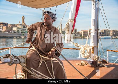 Membre de l'équipage tailler les voiles vêtues de vêtements traditionnels sur un bateau Dahabiya du Nil avec une ville égyptienne traditionnelle au loin Banque D'Images