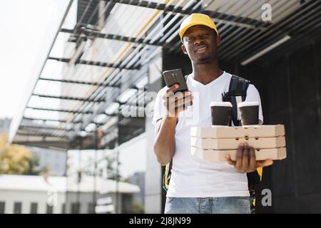 Service de livraison de nourriture dans un chapeau jaune contenant des boîtes à pizza et des tasses à café à emporter livrant la commande avec sac thermique sur son dos. Messagerie africaine avec téléphone à la main recherche adresse Banque D'Images