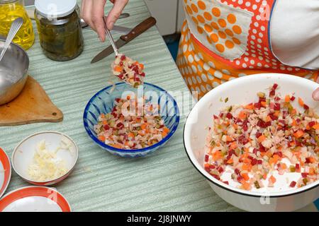 Gros plan de la table de cuisine avec les mains de la femme qui transfère la salade russe avec une cuillère du bol en émail blanc à un en plastique bleu clair. Banque D'Images