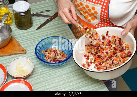 Gros plan de la table de cuisine avec les mains de la femme qui transfère la salade russe avec une cuillère du bol en émail blanc à un en plastique bleu clair. Banque D'Images