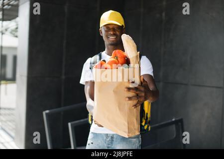 Concept de livraison. Messagerie afro-américaine avec un emballage contenant des produits frais du magasin. Un liveur africain souriant tenant un sac de papier avec des légumes biologiques. Banque D'Images