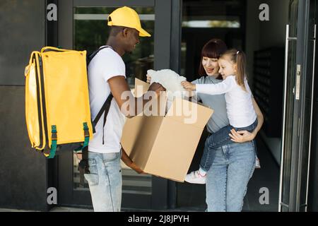 Belle jeune femme tenant sa fille dans les bras, ouvre les portes de sa maison et rencontre l'homme de livraison qui donne sa boîte en carton avec l'ours en peluche des services de commerce électronique. Banque D'Images