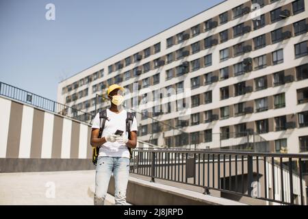 Un jeune coursier africain en masque de visage descend dans la rue avec sac à dos thermique, livrant la commande d'un restaurant ou d'un magasin. Le coursier parle au téléphone et prend la commande. Livraison rapide de divers produits. Banque D'Images