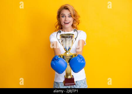 Femme joyeuse célébrant la victoire. Une femme en gants de boxe tient le trophée de la coupe gagnante du champion. Boxeur féminin primé isolé sur fond jaune. Banque D'Images