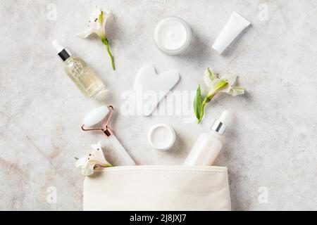 Sac à maquillage avec rouleau de visage de jade blanc, gua sha, hydratant, sérum, fleurs sur table de pierre. Concept de soin de la peau. Banque D'Images