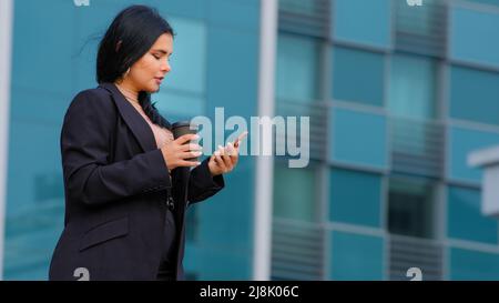 Jeune femme d'affaires hispanique marchant dans la rue arrière-plan du bâtiment de bureau vérifie e-mail sur le téléphone utilise l'application mobile rend l'achat en ligne envoie du texte Banque D'Images