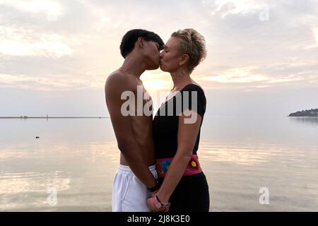 Couple multiculturel embrassant à côté de la mer pendant le coucher du soleil Banque D'Images