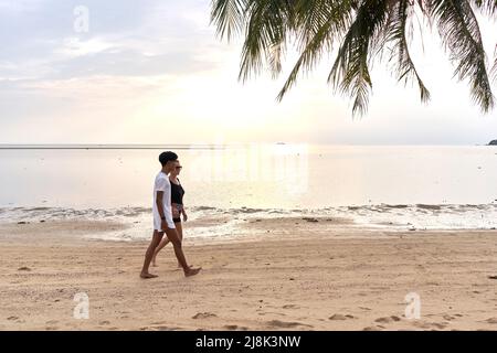 Photo panoramique d'un couple marchant le long d'une plage tropicale de sable au coucher du soleil Banque D'Images
